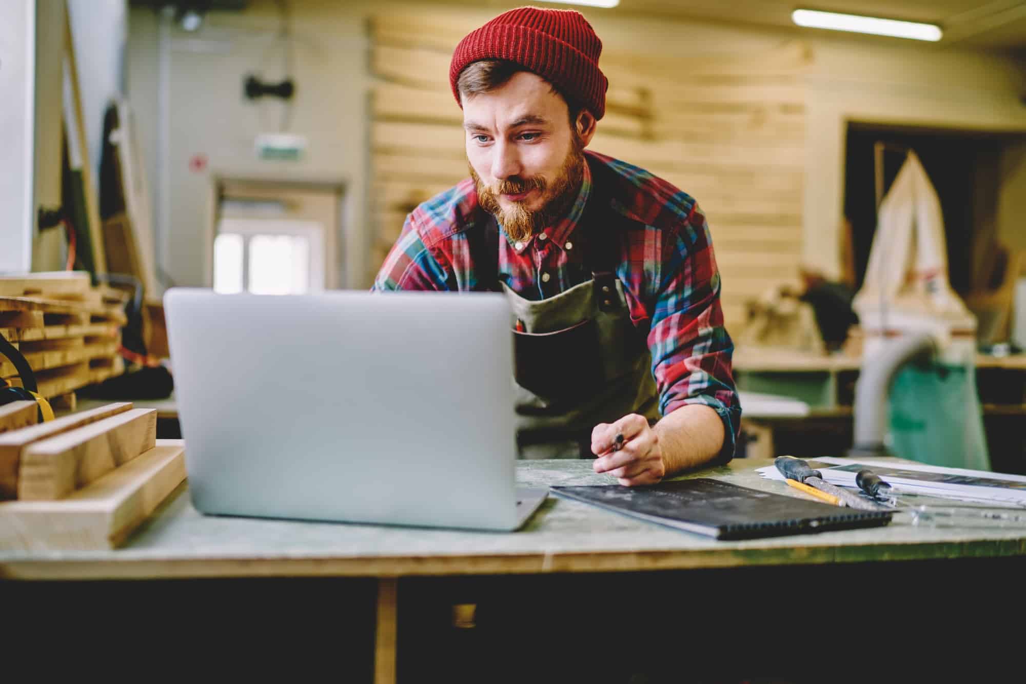 Creative bearded craftsman in apron working with laptop at workshop